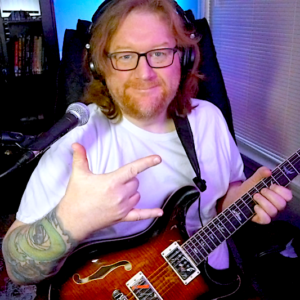 Me facing the camera with a closed mouth smile, right hand signing "I love you" while the other is holding the neck of the guitar in a sitting position. A microphone is off to the side, with a bookcase with closed glass doors behind him. Room is dim with purposeful lighting.